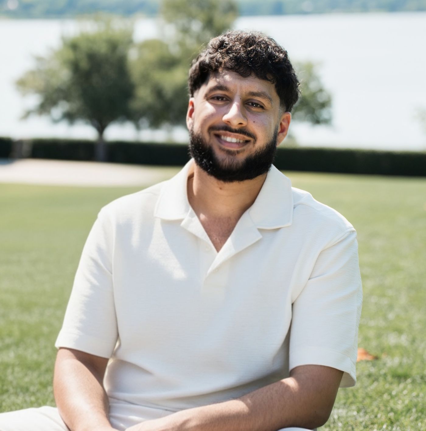 Rahim sitting outdoors on the grass by a lake, smiling at the camera.